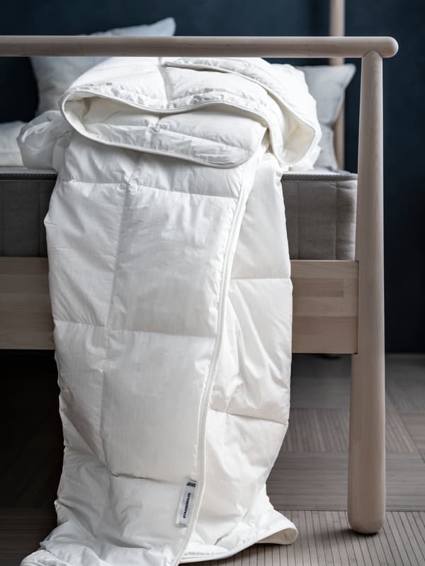 Duvet STRANDMOLKE tanpa sarung kuilt yang dilipat sembarangan terjuntai di sisi katil kayu birch GJÖRA di dalam bilik tidur bercahaya samar-samar.