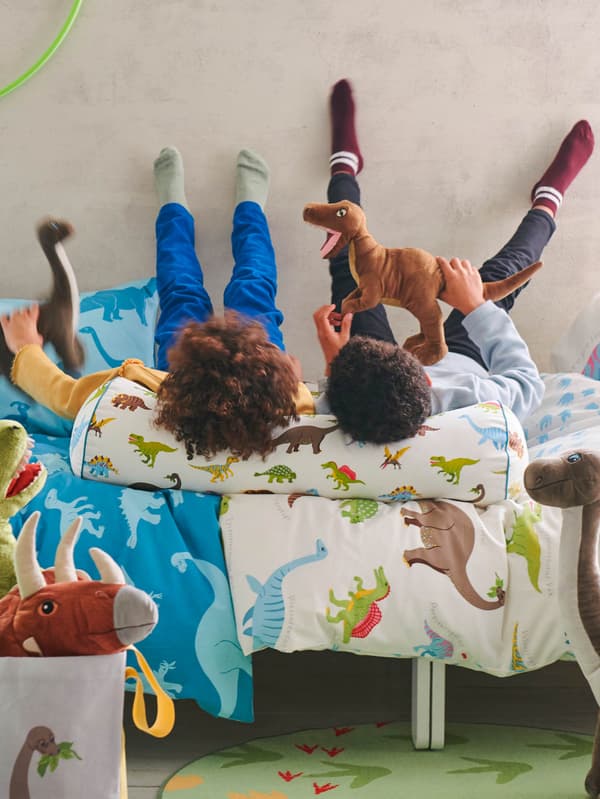 Dvě děti leží na posteli s povlečením JÄTTELIK, hlavy si opírají o polštář JÄTTELIK a hrají si s plyšovými dinosaury JÄTTELIK.