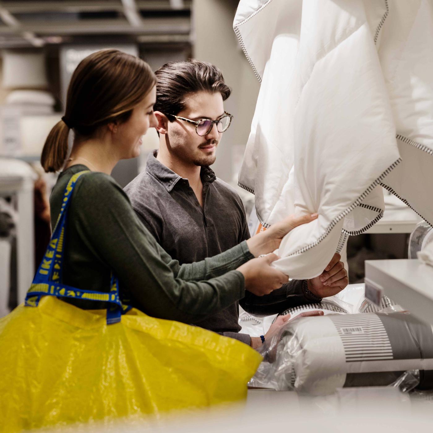 Dve osobe, jedna sa žutom IKEA torbom za kupovinu na ramenu, gledaju i dodiruju beli jorgan.