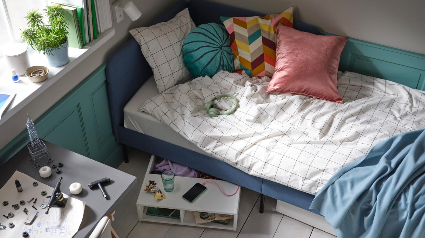 Een blauw gestoffeerd bed met wit/zwart geruit VITKLÖVER beddengoed en verschillende kussens, plus FREDVANG opberger voor onder het bed.