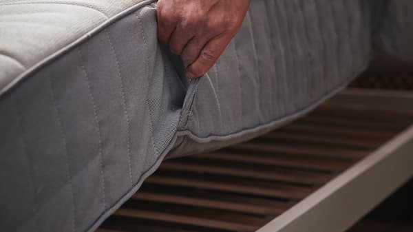 Een hand tilt een stevige/lichtblauwe VALEVÅG pocketveringmatras op van een bedframe.