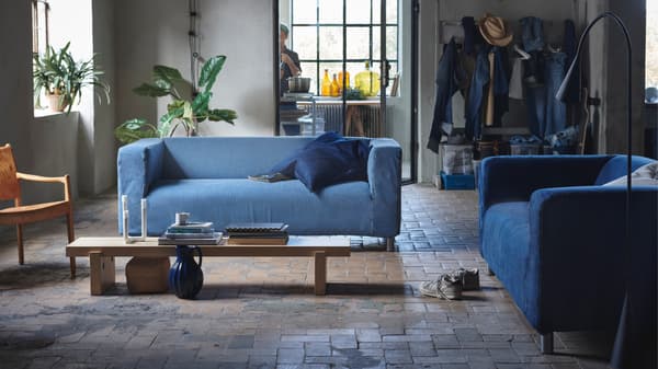 Een industrieelachtige loft met op een beige stenen vloer een houten lage lange rechthoekige salontafel met daarrond twee KLIPPANs met een hoes gemaakt van jeans.