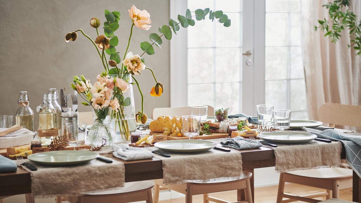 Een lange MÖRBYLÅNGA tafel feestelijk gedekt met lengtes van AINA stof, FÄRGKLAR borden, bloemen in vazen en diverse decoraties.