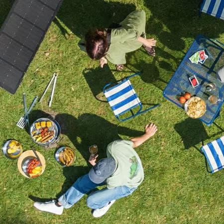 Een luchtfoto van twee mensen die picknicken op een weide. Om hen heen staan stoelen, een tafel, een parasol en draagbare zonnepanelen.