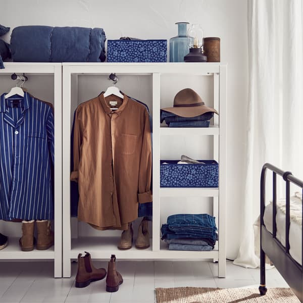 Een witte open IKEA HEMNES garderobekasten van massief hout, vol met blauwe en bruine kleding, hoeden en schoenen.