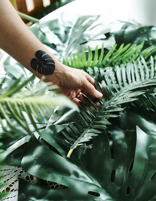Ein Arm mit einer Tätowierung in Form eines Blattes hält SMYCKA Kunstblatt Monstera/grün.