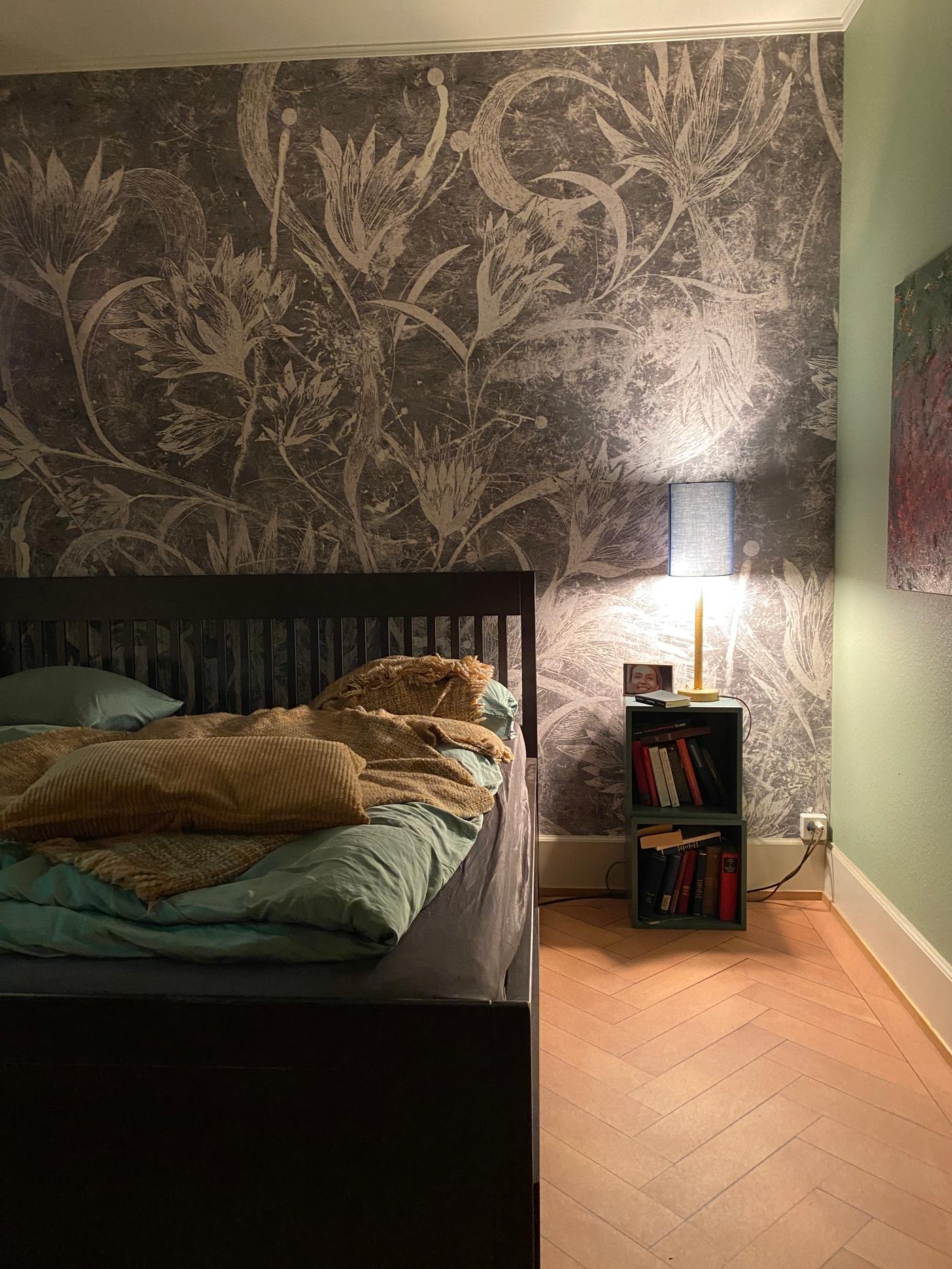 Ein Bett mit Bettwäsche darauf steht an einer Wand mit Fototapete mit floralem Muster. Daneben ein kleiner Nachttisch mit Büchern darin und einer Nachttischlampe darauf. 