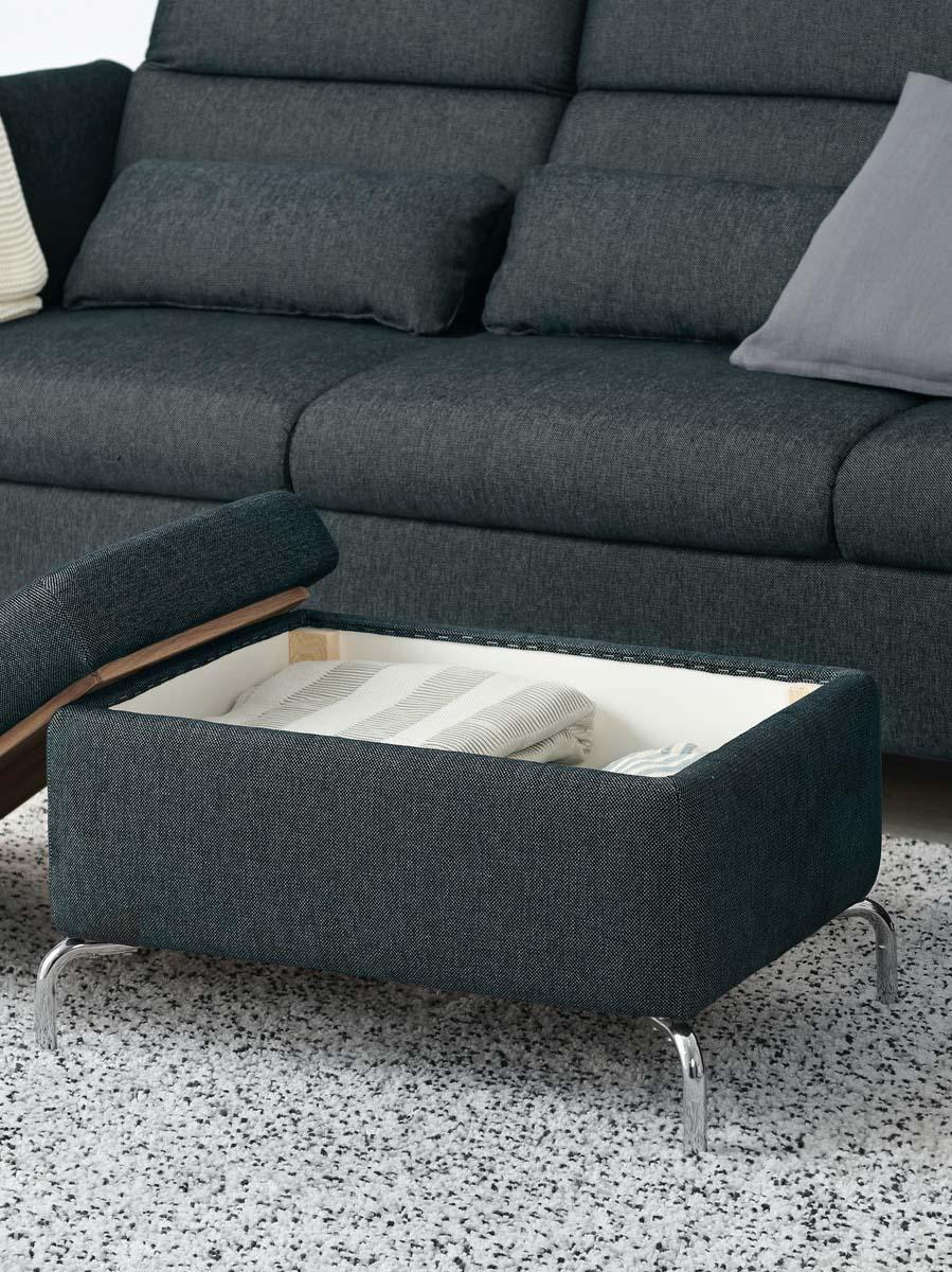 Ein dunkelgrauer FAMMARP Hocker mit silberfarbenen Beinen steht vor einem Sofa. Der abnehmbare Deckel des Hockers, der auch als Couchtisch verwendet werden kann, wenn man in wendet, ist seitlich angelehnt.