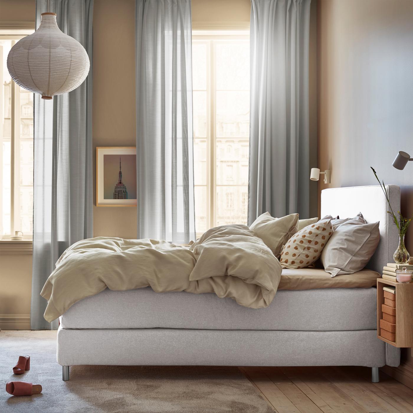 Ein DUNVIK Boxspringbett steht hier in einem hellen Schlafzimmer mit zwei grossen Fenstern. Auf dem Teppich am Fusse des Bettes liegen Schuhe.