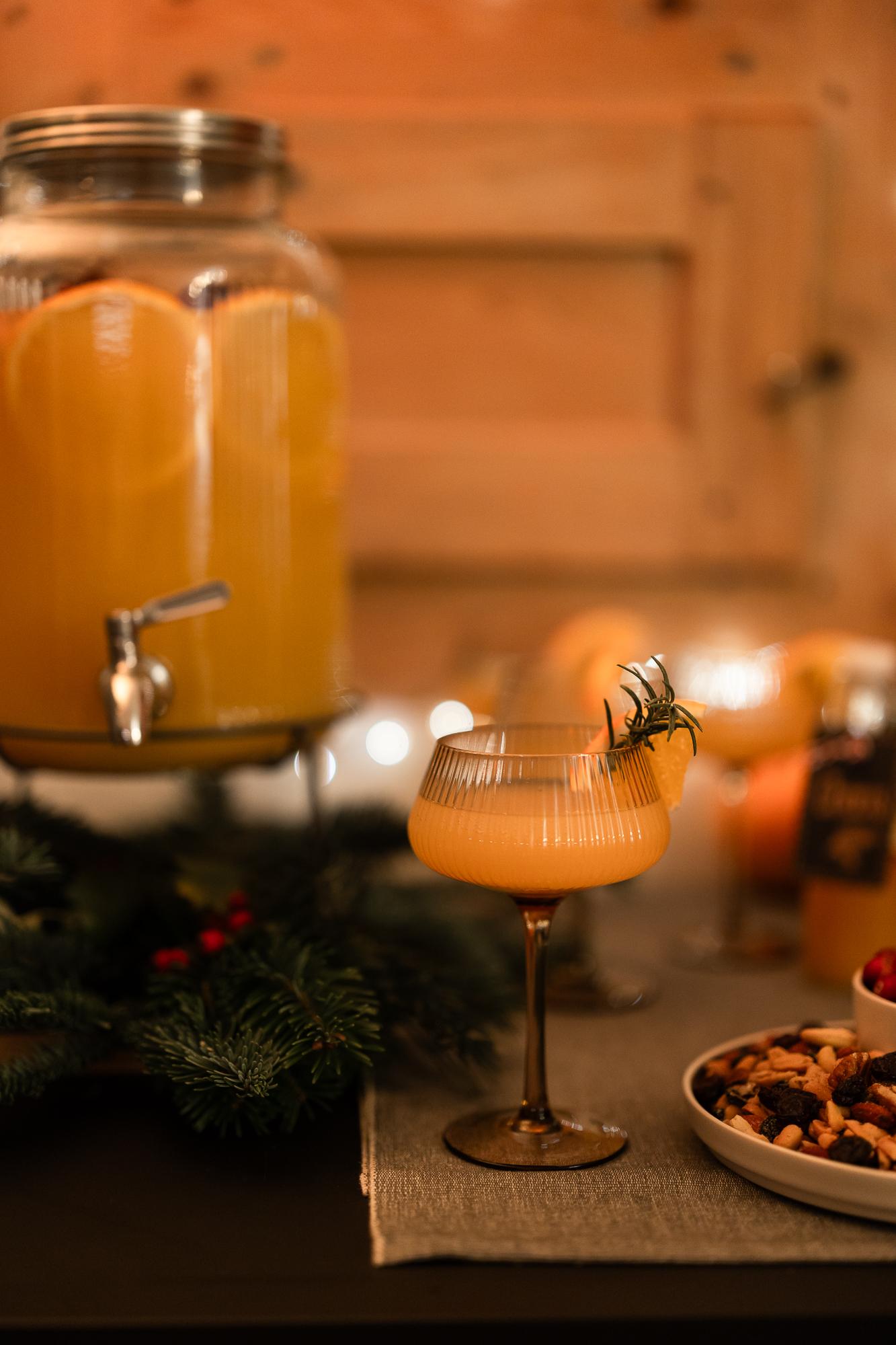 Ein festlicher Feiertagstisch mit einem Glas-Punschspender, gefüllt mit einem orangefarbenen Getränk, einem garnierten Cocktailglas, Nüssen und Grünpflanzen, beleuchtet von warmem Licht.
