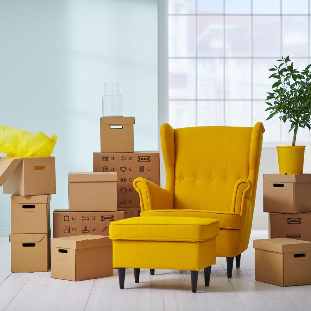 Ein gelber STRANDMON Sessel steht zwischen vielen IKEA Umzugsboxen.