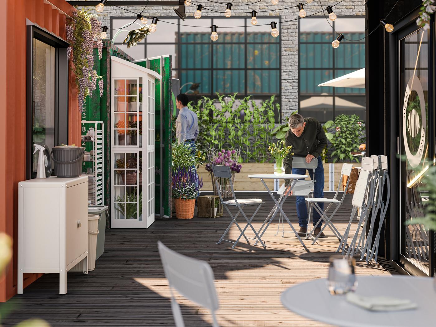 Ein grauer SUNDSÖ Stuhl wird von einer Person in einem schwarzen Hemd in einen Aussenbereich eines Cafés gestellt. Hier befinden sich ausserdem viele Pflanzen und ein Tisch mit weiteren Stühlen.