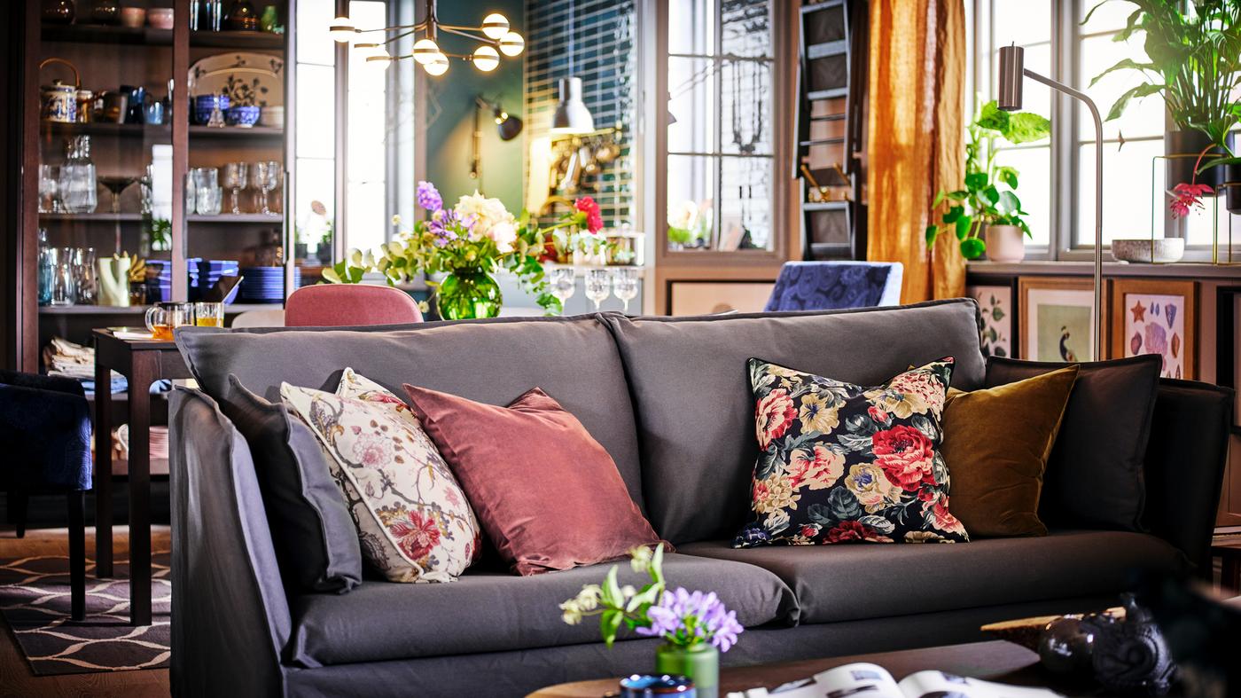 Ein graues BACKSÄLEN Sofa mit vier Dekokissen vor einem Esstisch mit Stühlen und einer Wand mit gerahmten Bildern dahinter.