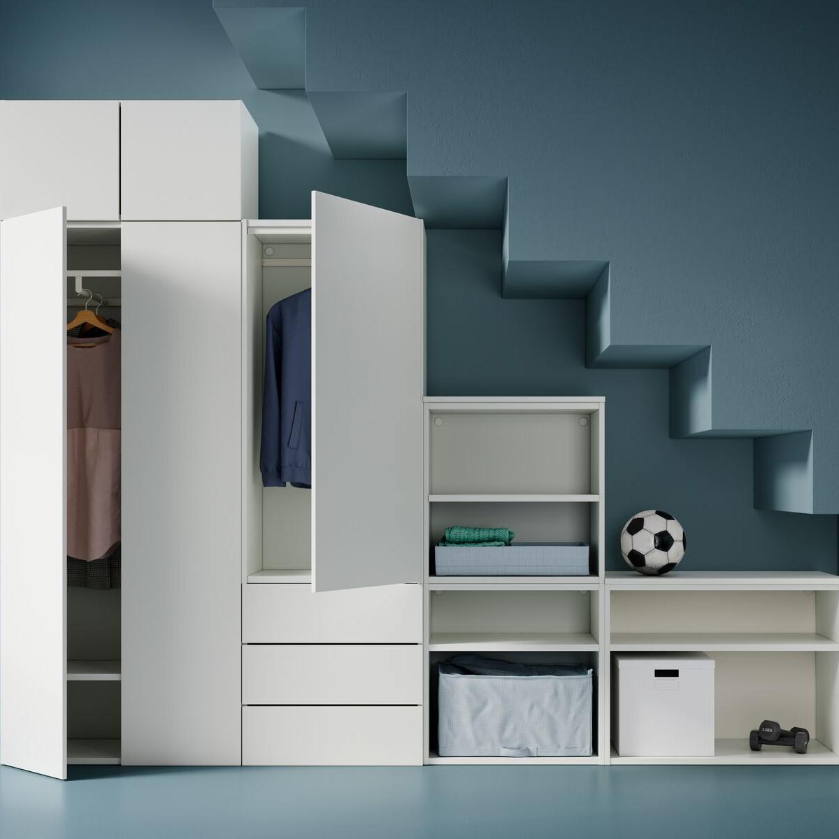 Ein großer PLATSA Kleiderschrank mit Elementen in unterschiedlichen Höhen unter einem angedeuteten Treppenaufgang. Darin und darauf sind Kleidung, Boxen und Fächer, Gewichte und ein Fußball zu sehen.