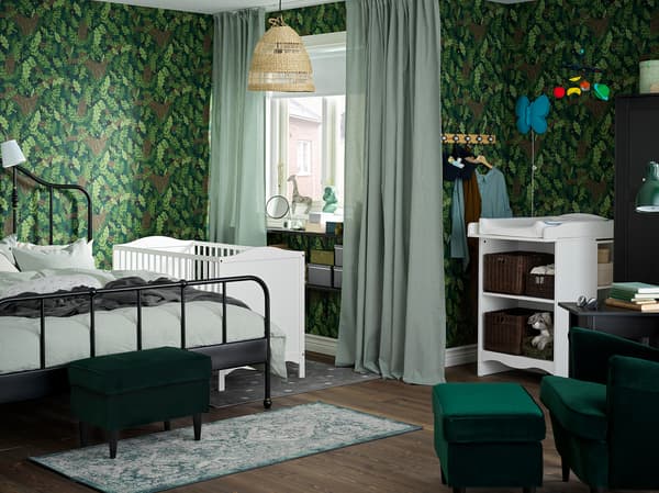 Ein grün gehaltenes Schlaf-/Kinderzimmer mit Wickeltisch und Babybett in Weiss und einem schwarzen Bettgestell für die Eltern.