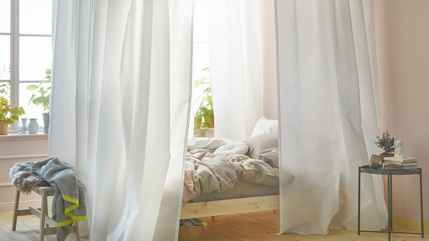 Ein helles Schlafzimmer mit einem Bett in Fensternähe. Das Bett hat einen Betthimmel, der aus weissen Gardinen und einer VIDGA Gardinenschiene gestaltet wurde.