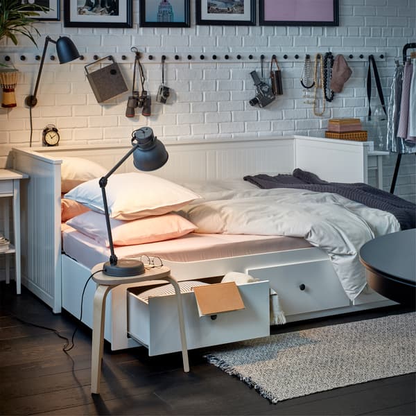 Ein HEMNES Tagesbett in Weiss mit drei Schubladen in seiner Bettfunktion. Neben dem Bett ist ein Hocker mit Leuchte zu sehen.
