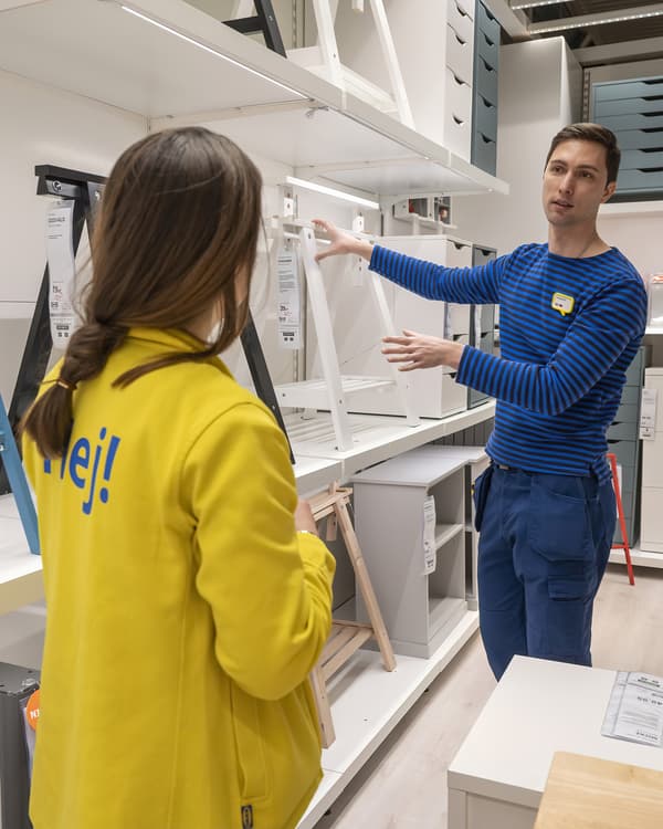 Ein IKEA-Mitarbeiter in blauem Streifenshirt erklärt einer Kollegin in gelber „Hej!“-Jacke verschiedene Tischböcke in einer Regalreihe der Ausstellung.