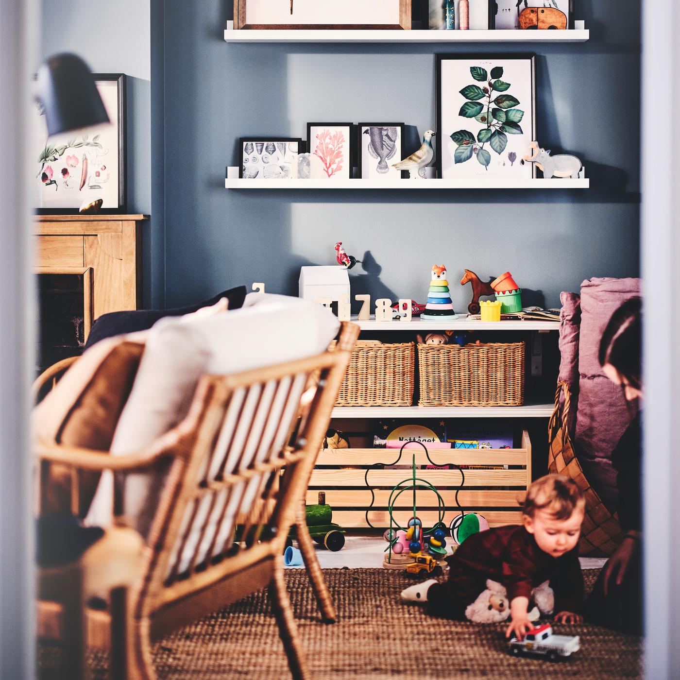 Ein Kind und ein Elternteil sind in einem Wohnzimmer mit weißen MOSSLANDA Bilderleisten, Rahmen, Rattankörben, Sesseln, einem Tisch und Spielzeug zu sehen.
