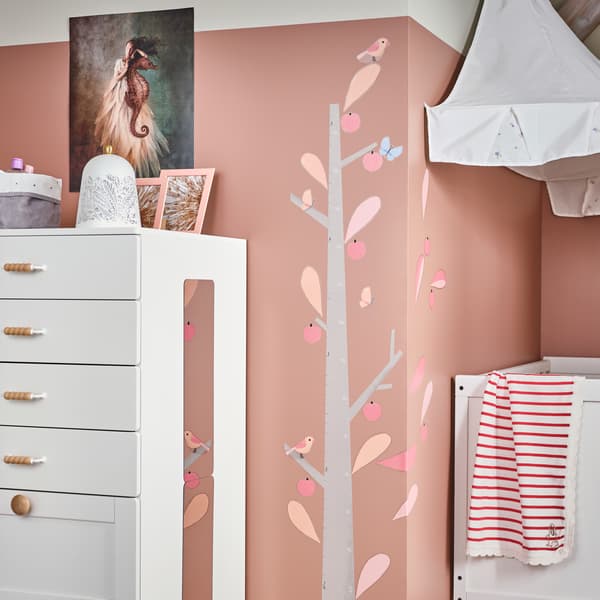 Ein Kinderzimmer mit einem Kleiderschrank mit einem Spiegel an einer Seite, einem weißen Betthimmel und Wänden in Rosa, an denen an einer Stelle Dekoaufkleber in Rosa/Weiß einen Baum bilden