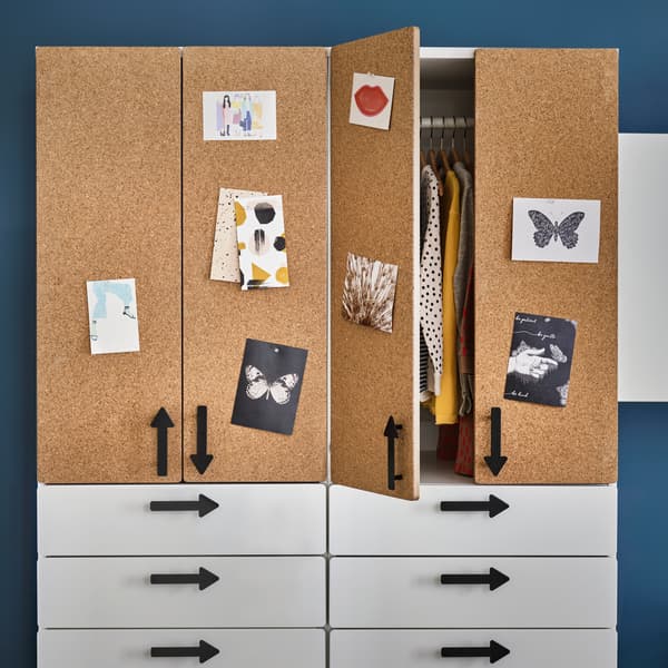 Ein Kleiderschrank mit vier weißen Schubladen, Türen aus Korkfurnier und Griffen in Pfeilform. Eine Tür steht offen, im Schrank hängt Kleidung an einer Kleiderstange.