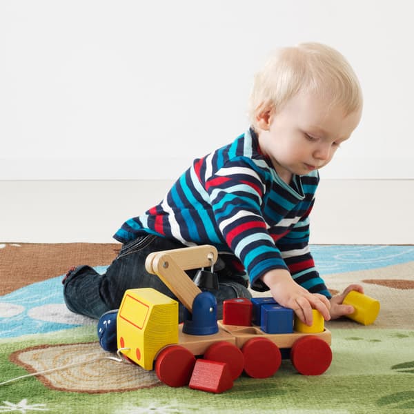 Ein Kleinkind spielt mit einem bunten IKEA MULA Kran mit Klötzen mit Bauklötzen auf einem blau-grünen Spielteppich.