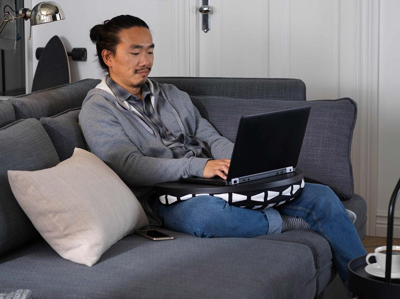 Ein Mann sitzt mit aufgeklapptem Laptop auf dem Schoss auf einem dunkelgrauen Sofa mit einem Beistelltisch davor auf dem eine Tasse Kaffee steht