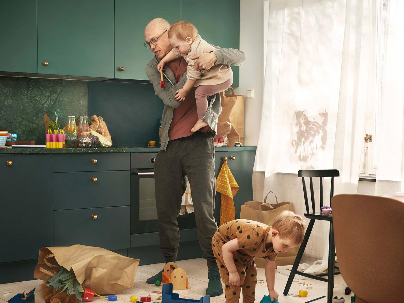 Ein Mann steht in einer Küche mit dunkelgrünen Fronten und hält ein Baby auf dem Arm. Er schaut erschrocken, weil die Einkaufstüte umgefallen ist. Ein Kleinkind hebt die Sachen vom Boden auf.