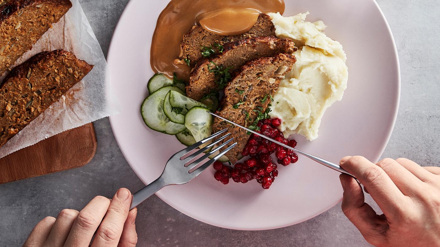 Ein Messer und eine Gabel werden an einen Teller mit Hackbraten, Gurkenscheiben, Preiselbeeren, Rahmsosse und Kartoffelpüree herangeführt.