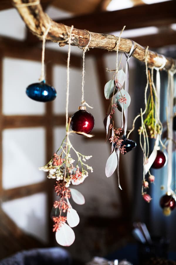 Ein mit Satinbändern, Weihnachtskugeln, Eukalyptus und Trockenblumen geschmückter Zweig, der über einem Esstisch reicht.