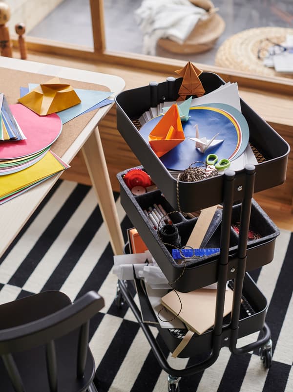 Ein RÅSKOG Servierwagen in Schwarz mit Bastelutensilien steht neben einem Tisch mit buntem Papier.