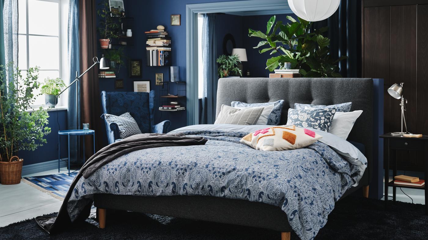 Ein Schlafzimmer in verschiedenen Blautönen, u. a. mit einem gepolsterten Bett, jeder Menge Kissen, einem Ohrensessel und einem grossen Teppich.