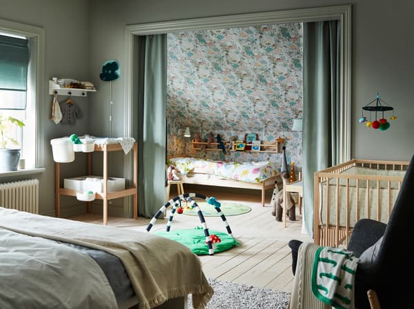 Ein Schlafzimmer mit einem Doppelbett, einem Wickeltisch und Babyaccessoires. Ein SNIGLAR Juniorbettgestell steht in einer Nische hinter einem grünen Vorhang.