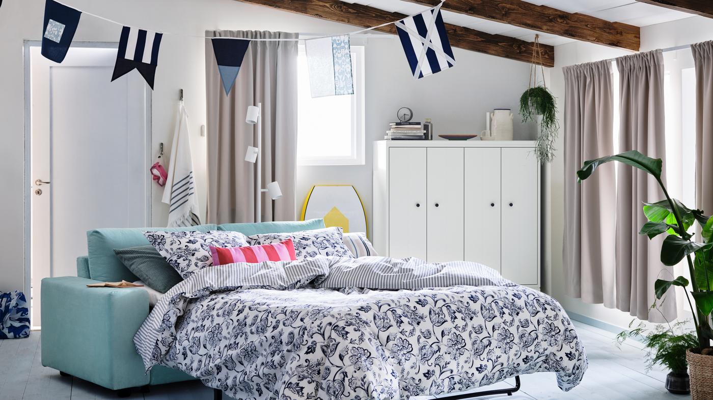Ein Sommerhaus mit Fenstern, an denen Gardinen mit Verdunklungsfunktion hängen, u. a. mit einem VIMLE 2er-Bettsofa mit der JUNIMAGNOLIA Bettwäsche.