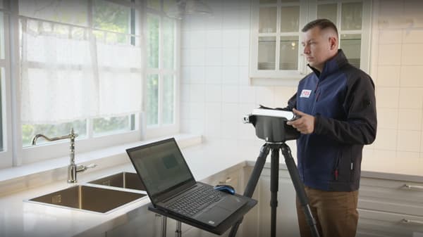 Ein Techniker vermisst eine moderne IKEA-Küche mit einem 3D-Laserscanner auf Stativ. Neben ihm ein geöffneter Laptop zur digitalen Küchenplanung, im Hintergrund eine Doppelspüle vor großen Fenstern mit Tageslicht