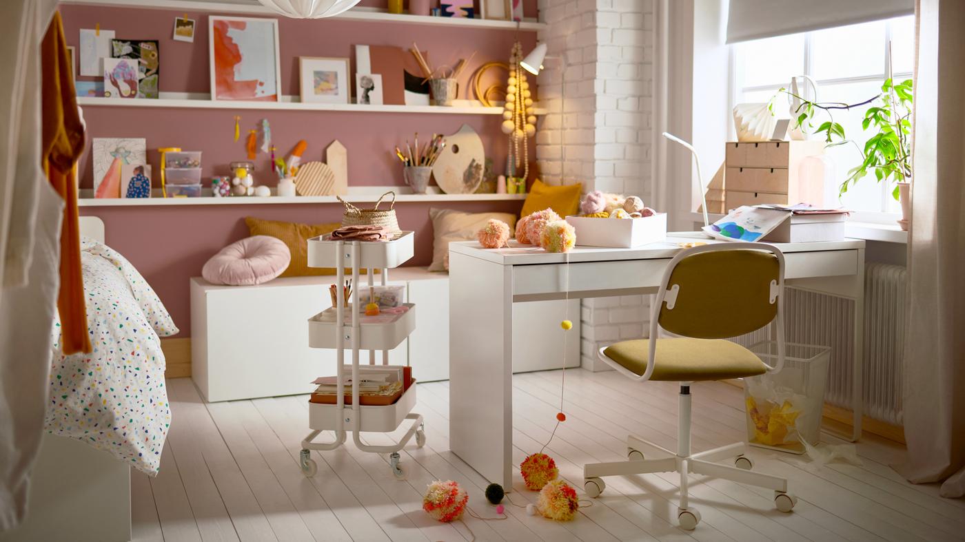 Ein Teenagerzimmer mit einem Schreibtisch, Rollwagen, Bilderleisten mit Deko, Kissen und einem Drehstuhl.