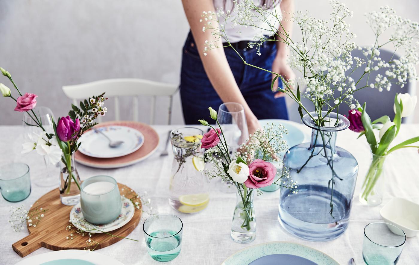 Ein Tisch mit pastellfarbenem Geschirr, frischen Blumen in Glasvasen und einem Holzbrett auf einer weissen Tischdecke.