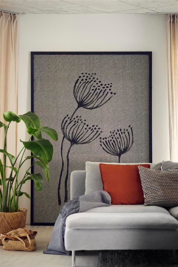 Ein übergrosses Bild aus Stoff mit schwarzem Rahmen und Blumen-Motiv steht auf dem Boden an der Wand hinter einem Sofa