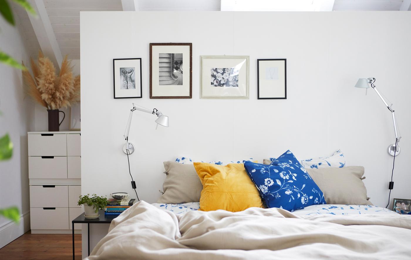 Ein weisses Schlafzimmer mit neutraler und geblümter Bettwäsche. An der Wand sind Fotos in verschiedenen Rahmen.