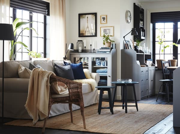 Ein Wohnzimmer mit einem hellbeigen Bettsofa, einem Juteteppich, einem Rattansessel, zwei Schränken und einer Kommode in Grau.
