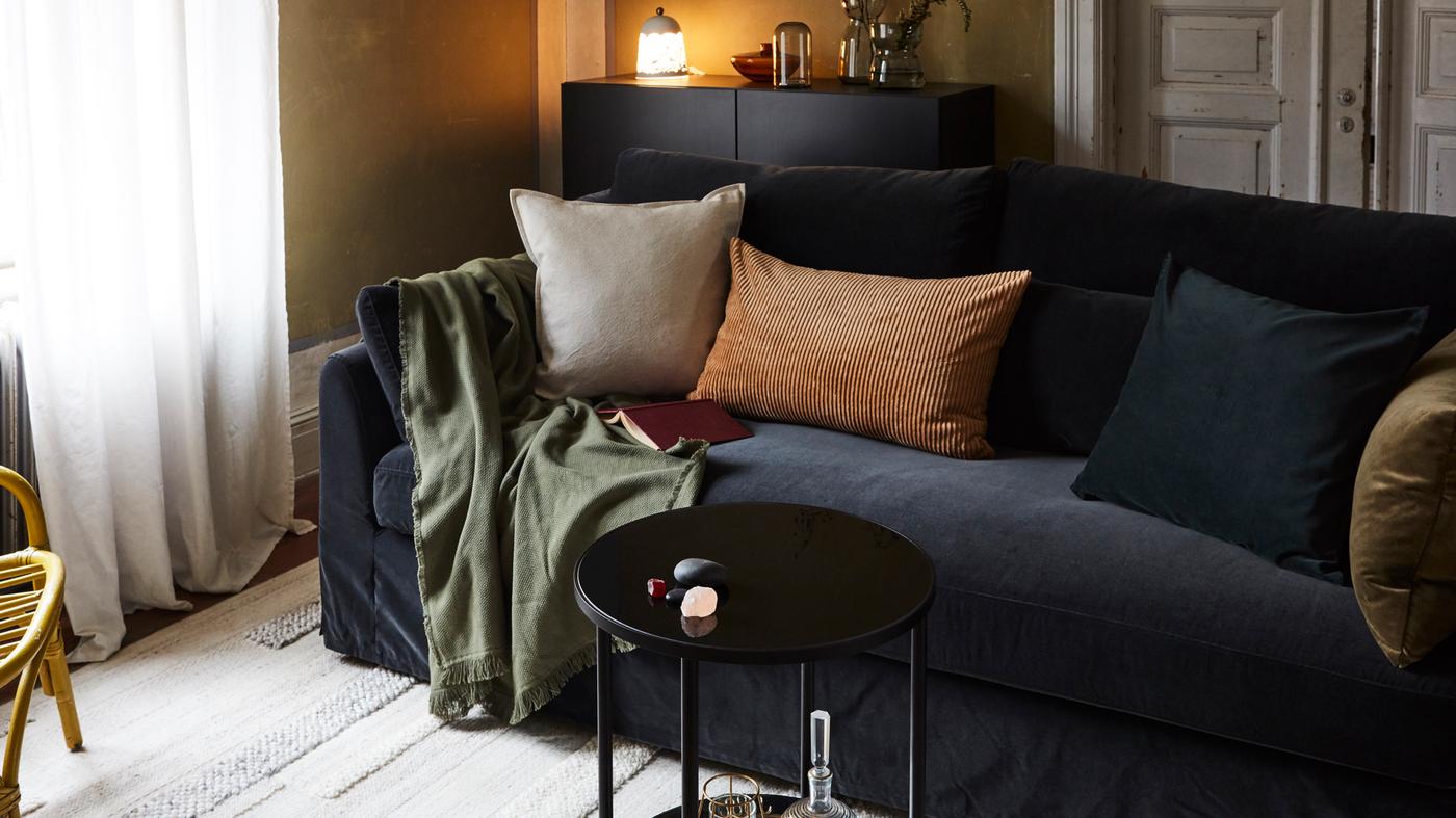 Ein Wohnzimmer mit einem Sofa, Kissen, einem Plaid, einem Teppich, einem kleinen Couchtisch, einem Sideboard mit einer Leuchte und Vasen darauf und ein Sessel aus Rattan.