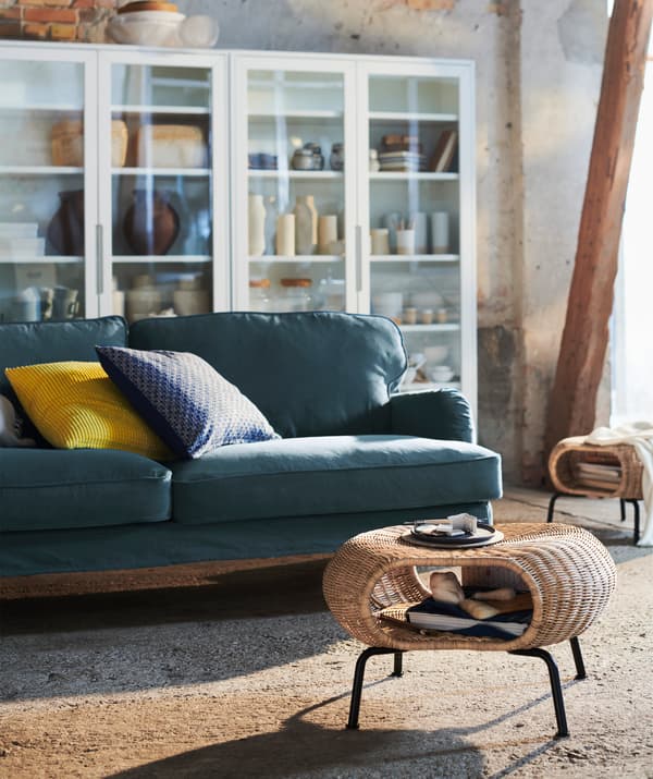 Ein Wohnzimmer mit GAMLEHULT Hocker mit Aufbewahrung Rattan/anthrazit, einem dunkelgrünen Sofa und einem Vitrinenschrank