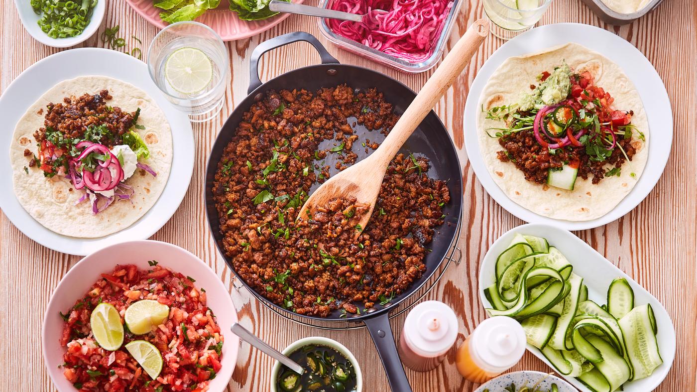 Eine Bratpfanne mit dem pflanzlichen Hack, umgeben von Schüsseln mit Pico de Gallo und Tortillas.
