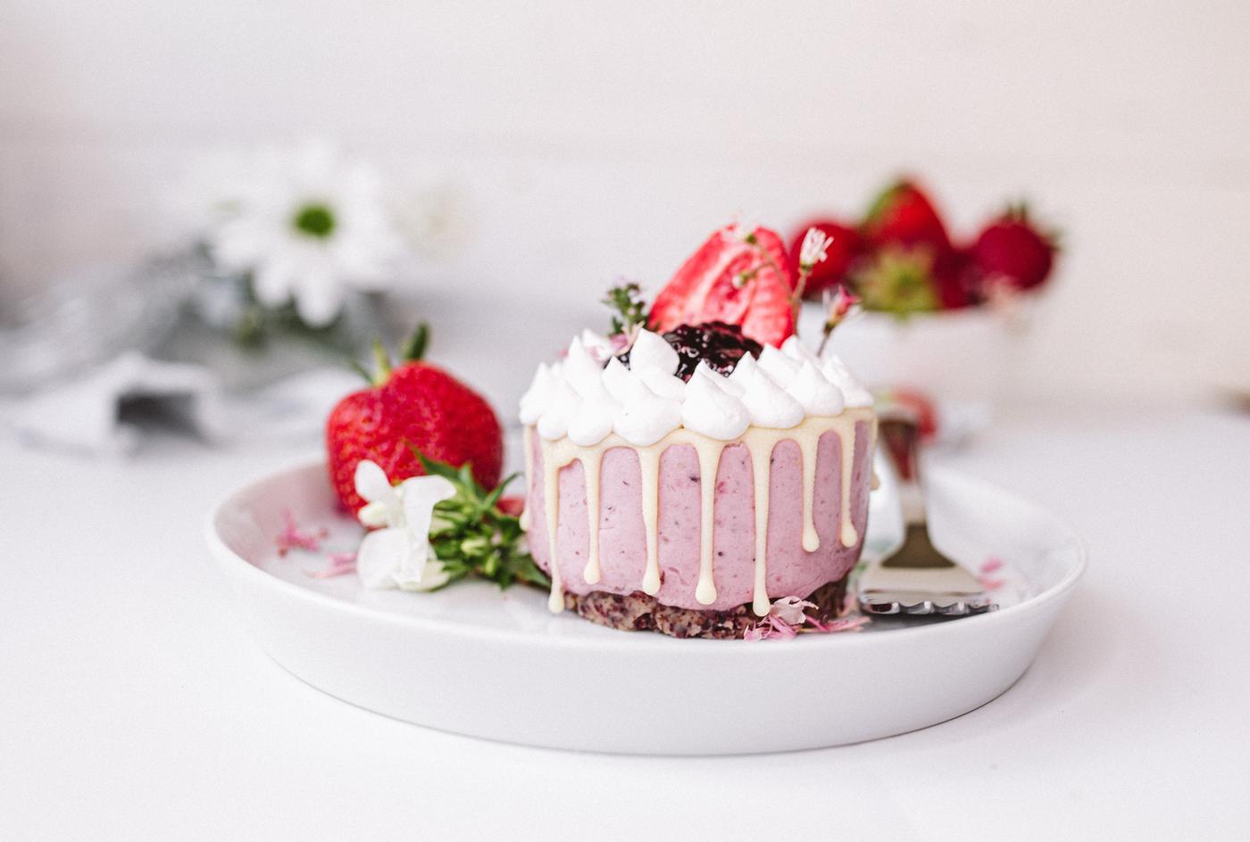Eine einzelne Portion Erdbeerkuchen, gekrönt mit Schlagsahne und einer frischen Erdbeere, auf einem weissen Teller mit einer Gabel, vor einem unscharfen Hintergrund.