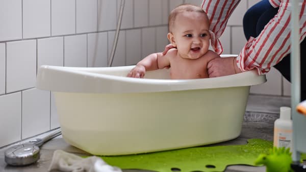 Eine erwachsene Person in einem rot-weiß gestreiften Hemd hält ein Baby in einer LÄTTSAM Babybadewanne fest. Vor der Wanne liegt eine grüne PATRULL Badewannenmatte.