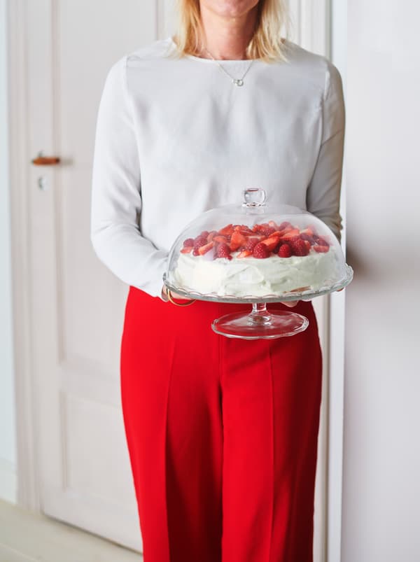 Eine Frau, die eine ARV BRÖLLOP Platte auf Fuss mit einem Kuchen darauf in einen Raum trägt.
