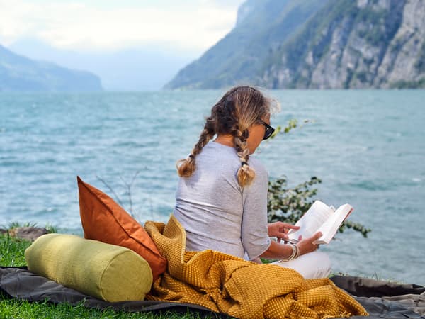 Eine Frau liest ein Buch, während sie auf einer senffarbenen Decke am See sitzt.