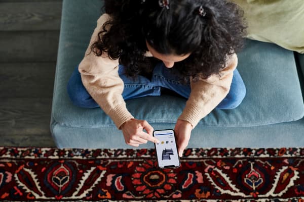 Eine Frau sitzt mit gekreuzten Beinen auf einem Hocker und überprüft die Produktverfügbarkeit für einen Sessel in der IKEA App. 
