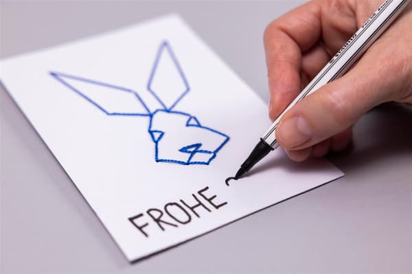 Eine Frau sticht mit einem Faden durch ein Paier und schreibt auf einen Zettel Frohe Ostern