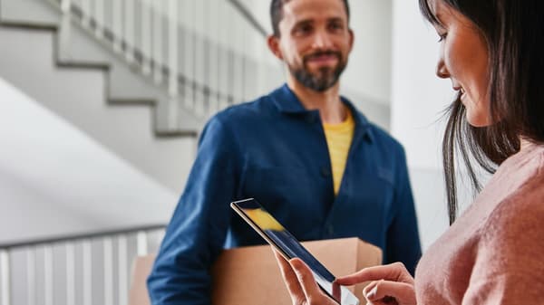 Eine Frau überprüft auf dem Smartphone die Details ihrer Paketlieferung während der Zusteller mit dem Paket lächelnd wartet